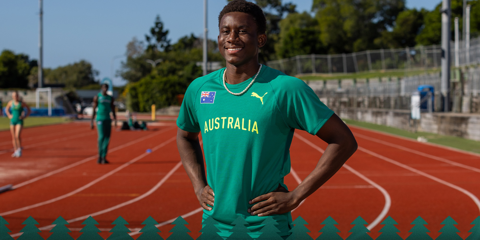 Smiling young athlete in green Australia track uniform stands on a red running track with hands on hips, teammates blurred in the background.