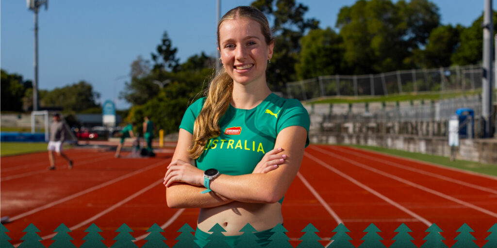 Smiling female track athlete in a green Australia jersey stands with arms crossed on an outdoor running track, stadium seating behind her.