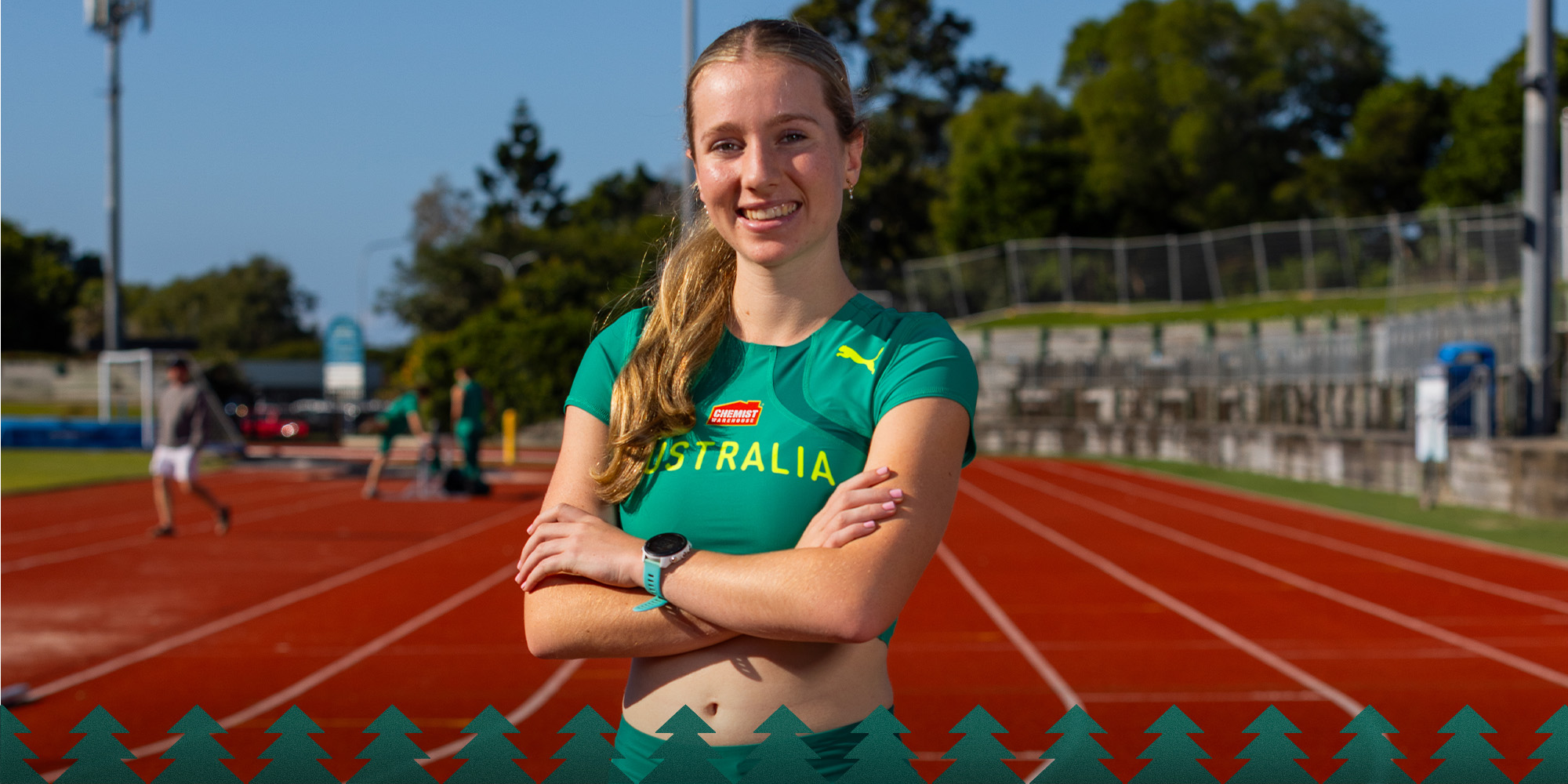 Smiling female track athlete in a green Australia jersey stands with arms crossed on an outdoor running track, stadium seating behind her.
