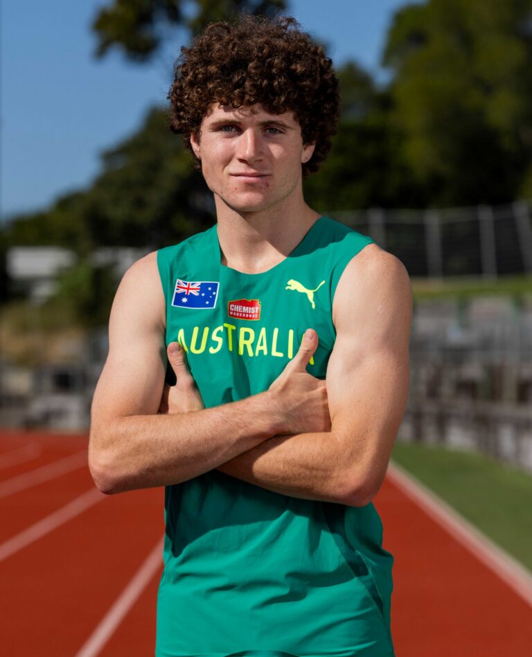Young male athlete in green Australia uniform posing with arms crossed on a running track outdoors.