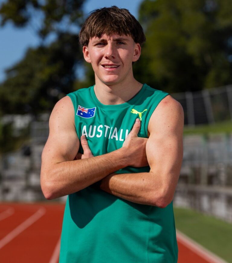 Young man in a teal Australia track jersey with 'AUSTRALIA' text stands on a running track, arms crossed, smiling.