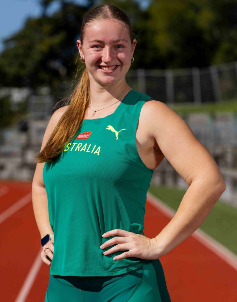 Smiling woman in a green athletic top standing on a red running track with hands on hips.