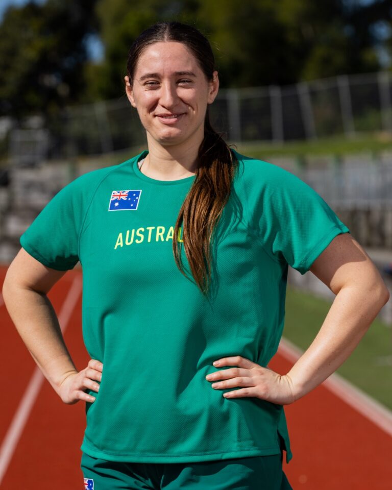 Female athlete standing on a running track with hands on hips, wearing a teal Australia jersey with a small Australian flag patch.