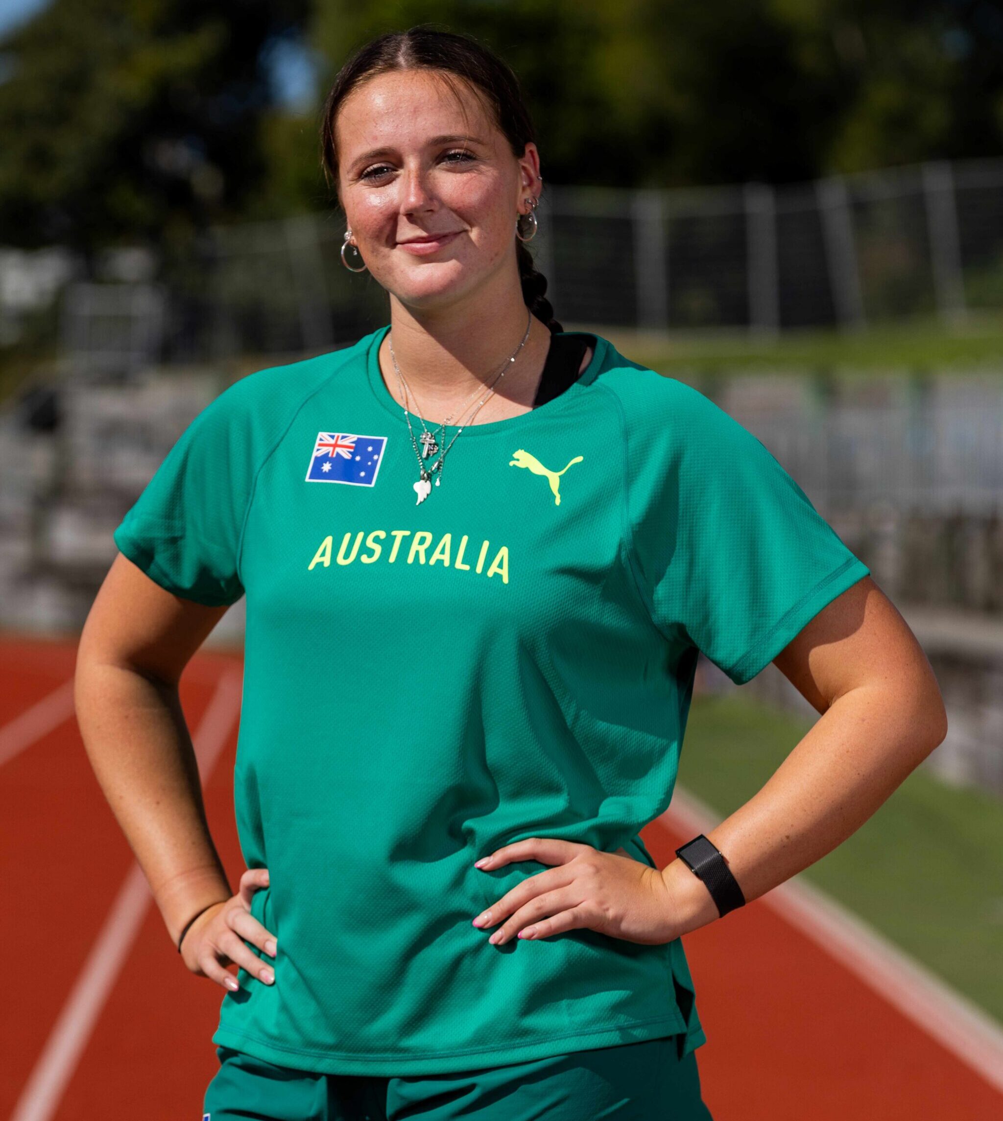 Smiling person in a green Australia sports shirt standing on a running track with hands on hips.