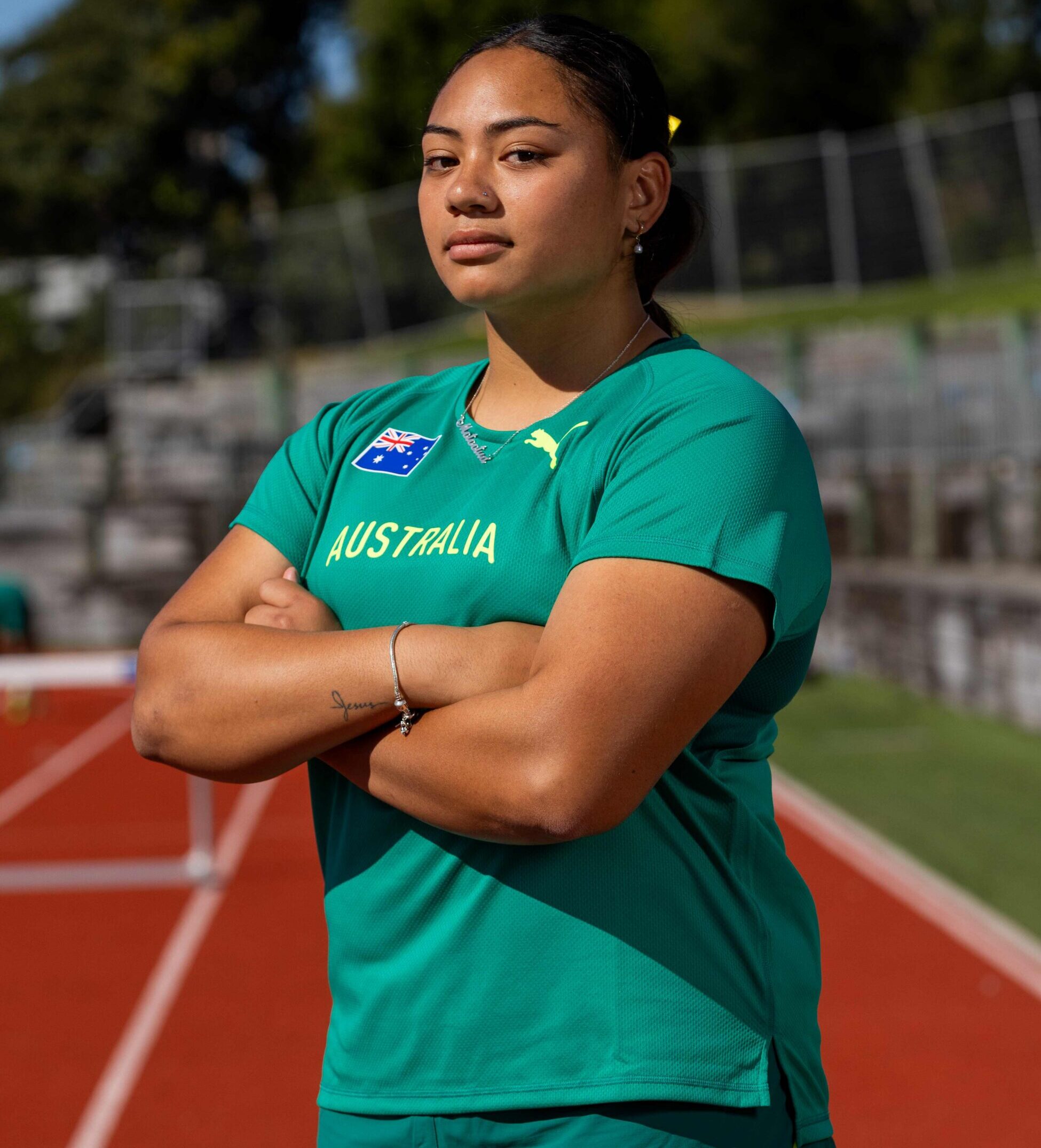 Athlete in teal Australia sports jersey stands on a running track with arms crossed, stadium seating in background.