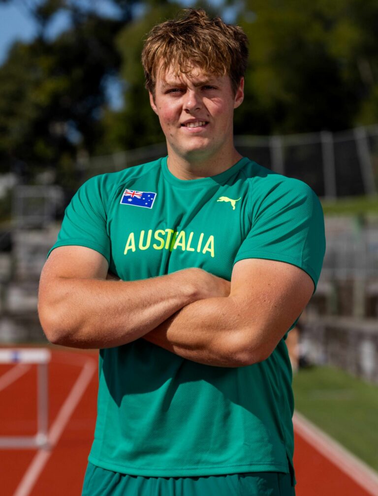 Athlete on a running track with arms crossed, wearing a teal Australia jersey with a small flag patch on the chest.
