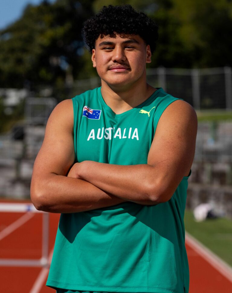 Athlete with curly black hair wearing a teal Australia singlet, arms crossed in front of a running track with a fence in the background