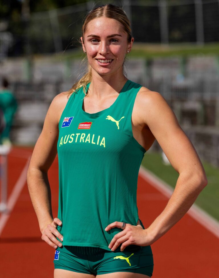 Australian female athlete in a green uniform standing on a running track, smiling with hands on hips; Australia team emblem visible on jersey.