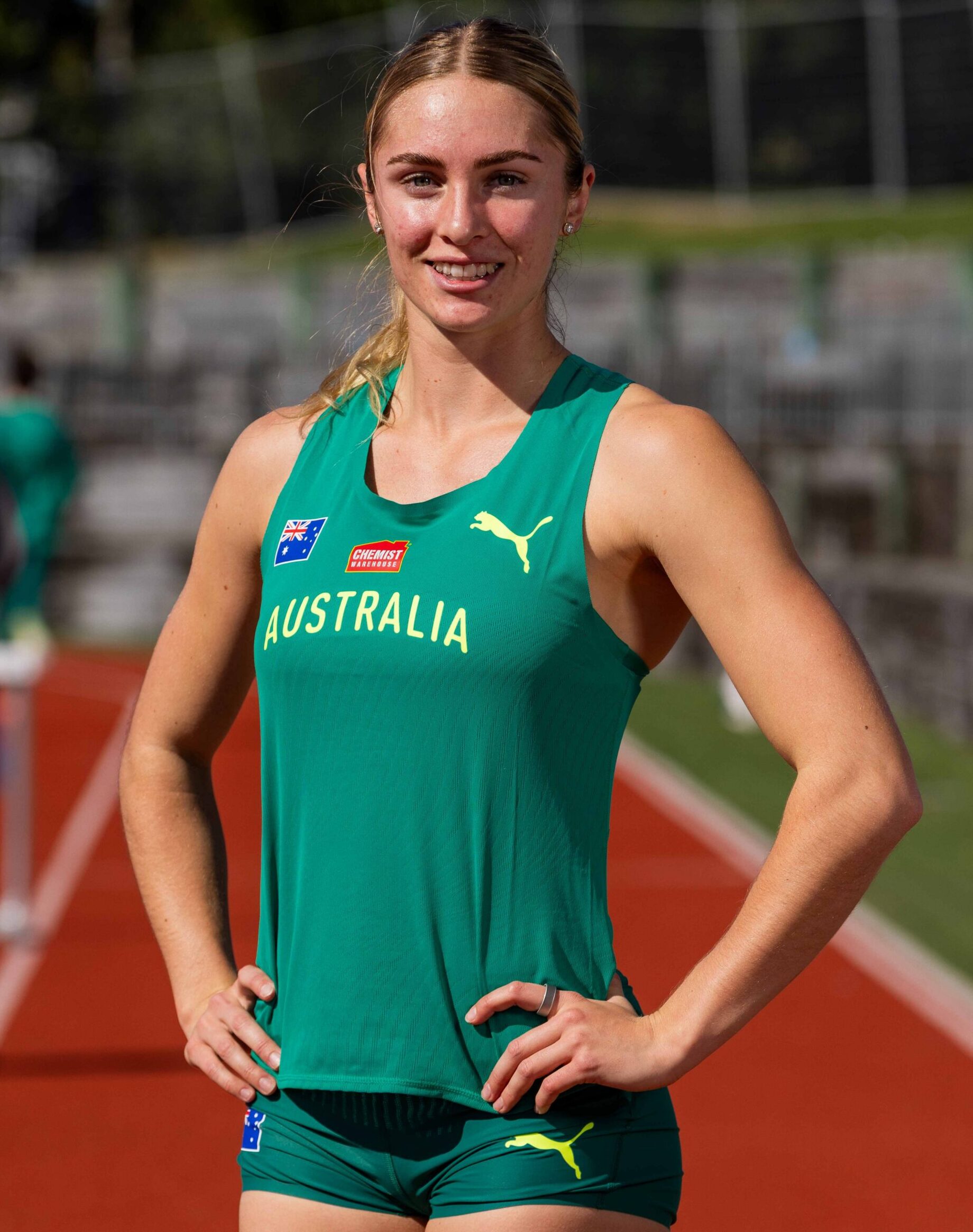 Australian female athlete in a green uniform standing on a running track, smiling with hands on hips; Australia team emblem visible on jersey.