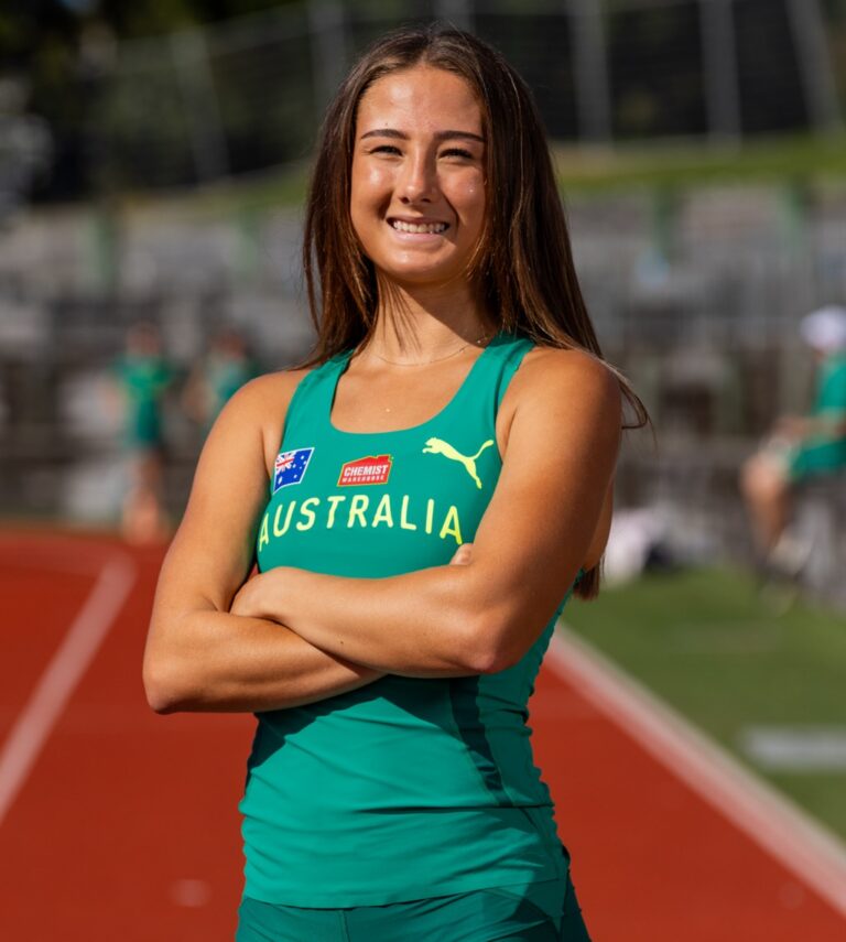 Australian female athlete standing with arms crossed on a running track, smiling, wearing a green Australia singlet with team logos.