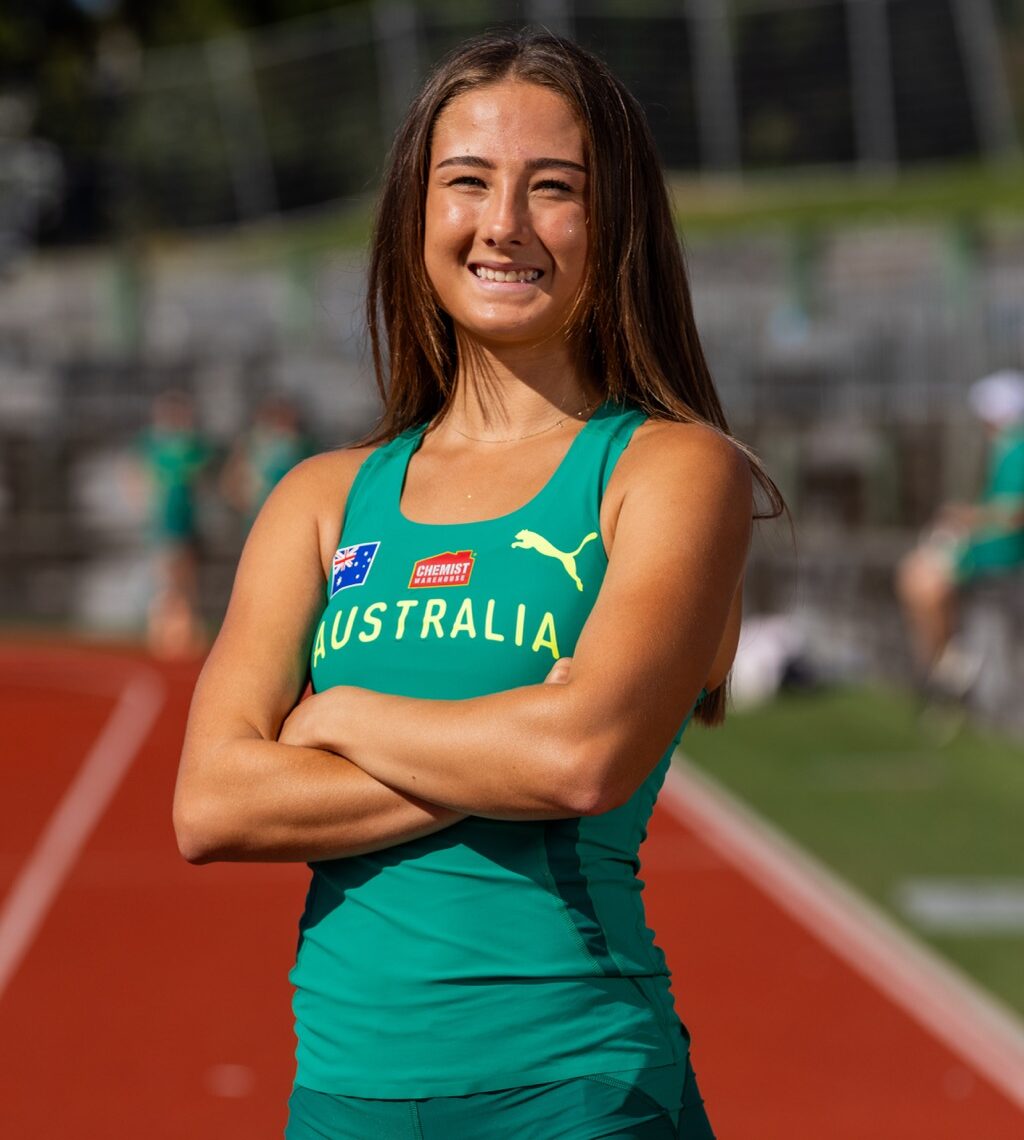 Australian female athlete standing with arms crossed on a running track, smiling, wearing a green Australia singlet with team logos.