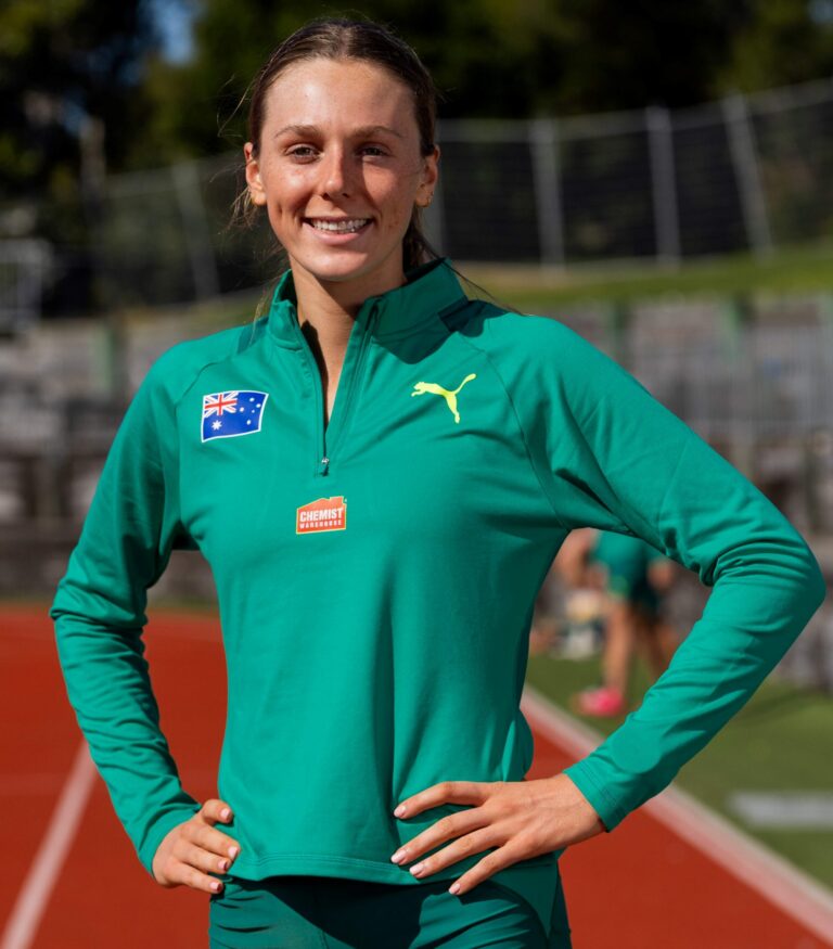 Smiling female athlete in a green track jacket with Australian flag patch on the chest, standing on a running track with hands on hips.