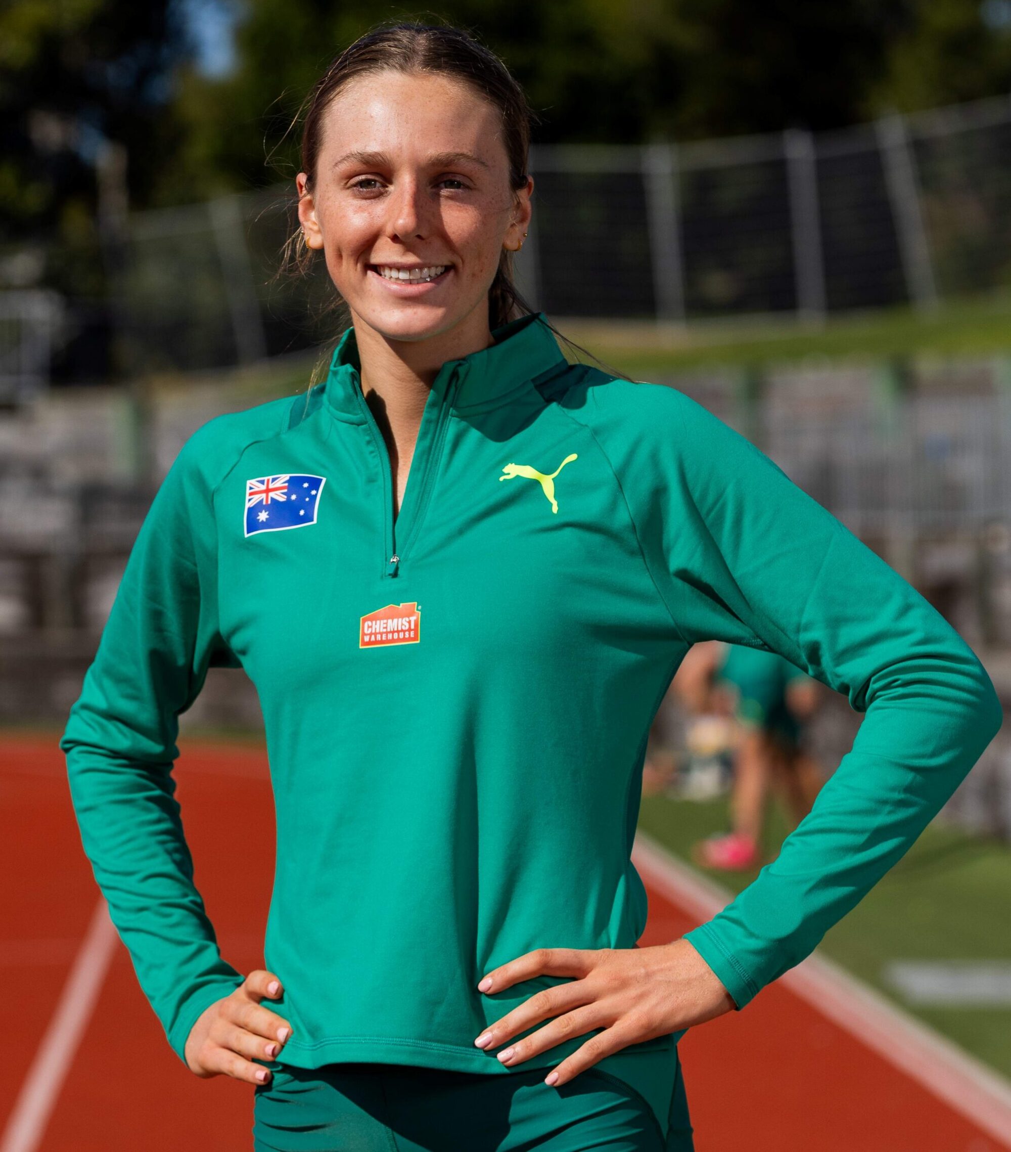 Smiling female athlete in a green track jacket with Australian flag patch on the chest, standing on a running track with hands on hips.