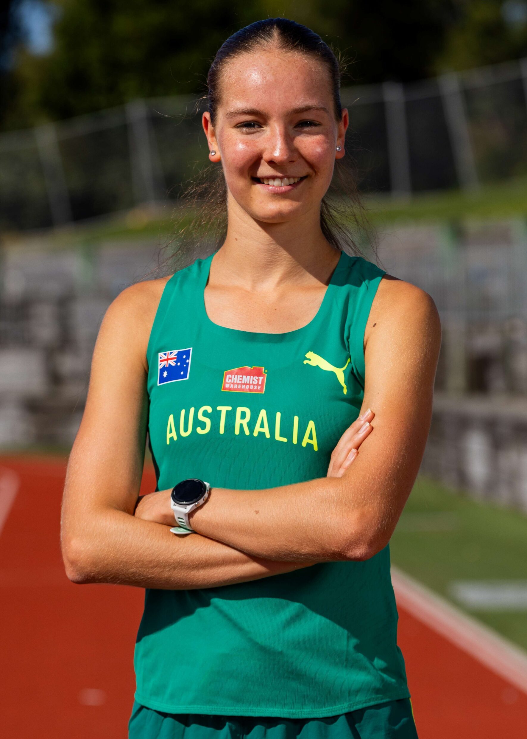 Smiling female athlete in a green Australia jersey stands on a running track with arms crossed.