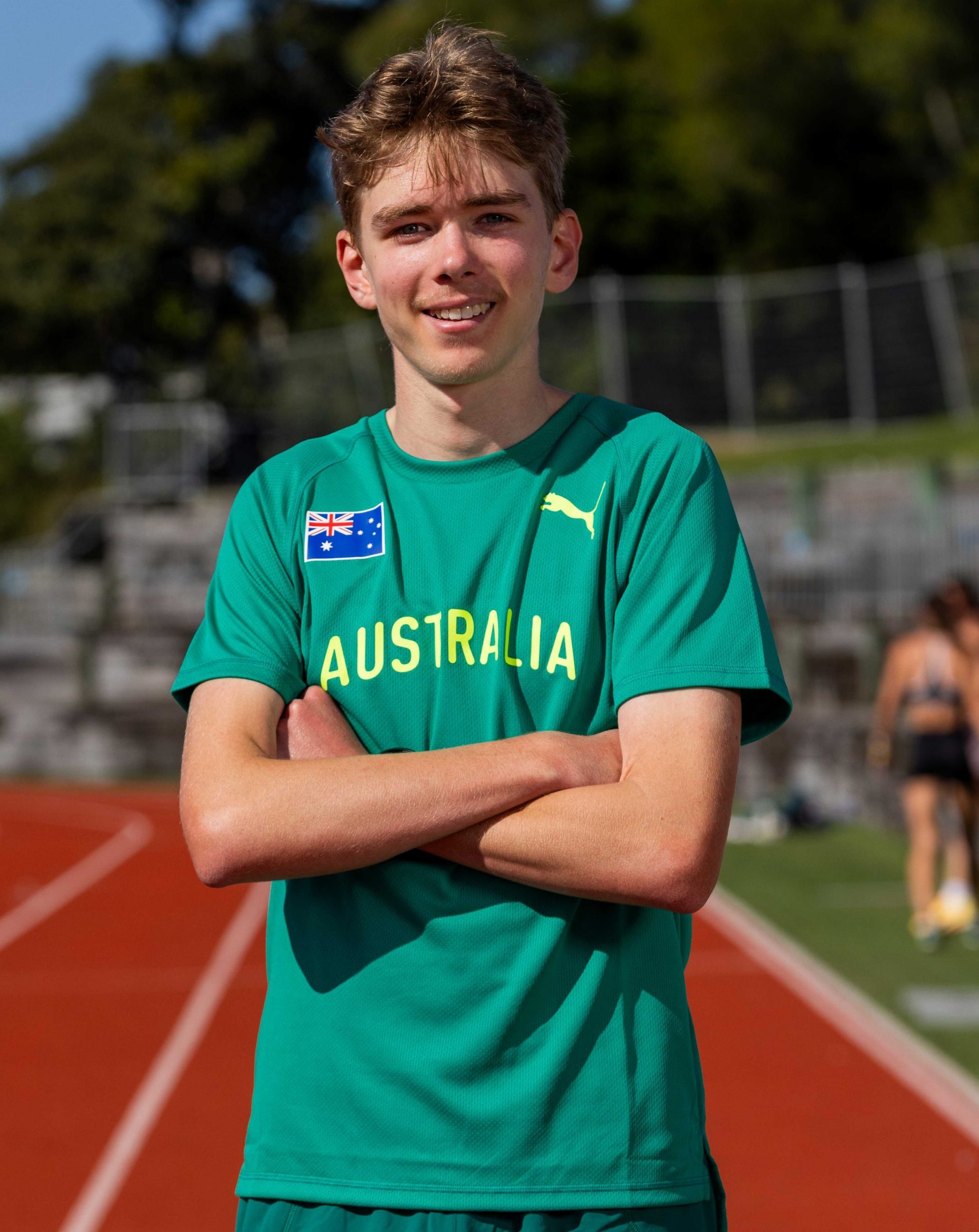 Smiling young man in a green Australia sports shirt standing on a running track with arms crossed.