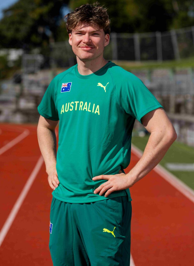 Male athlete in a green Australia jersey posing on a running track, hands on hips, outdoors.