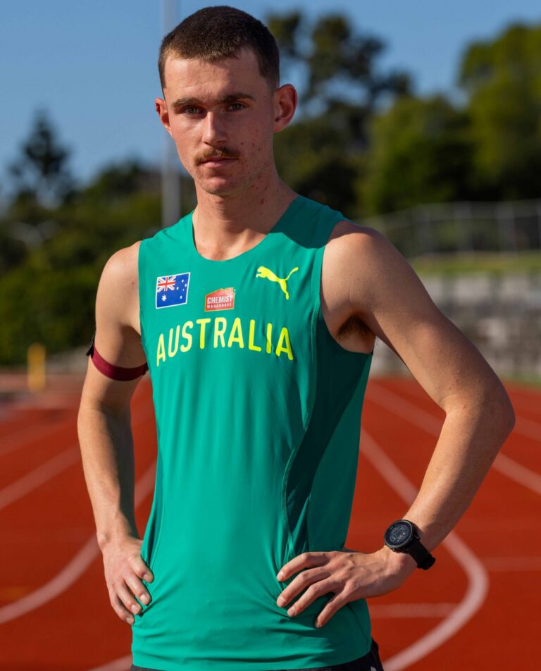 Male athlete in teal Australia jersey stands on a running track with hands on hips, blue sky in background.