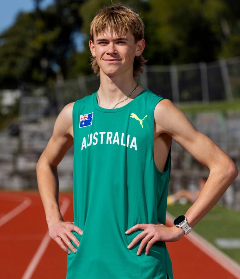 Young male athlete standing on a track with hands on hips, wearing a teal Australia uniform and a Newcastle/flag badge visible on the chest, outdoors on a sunny day.