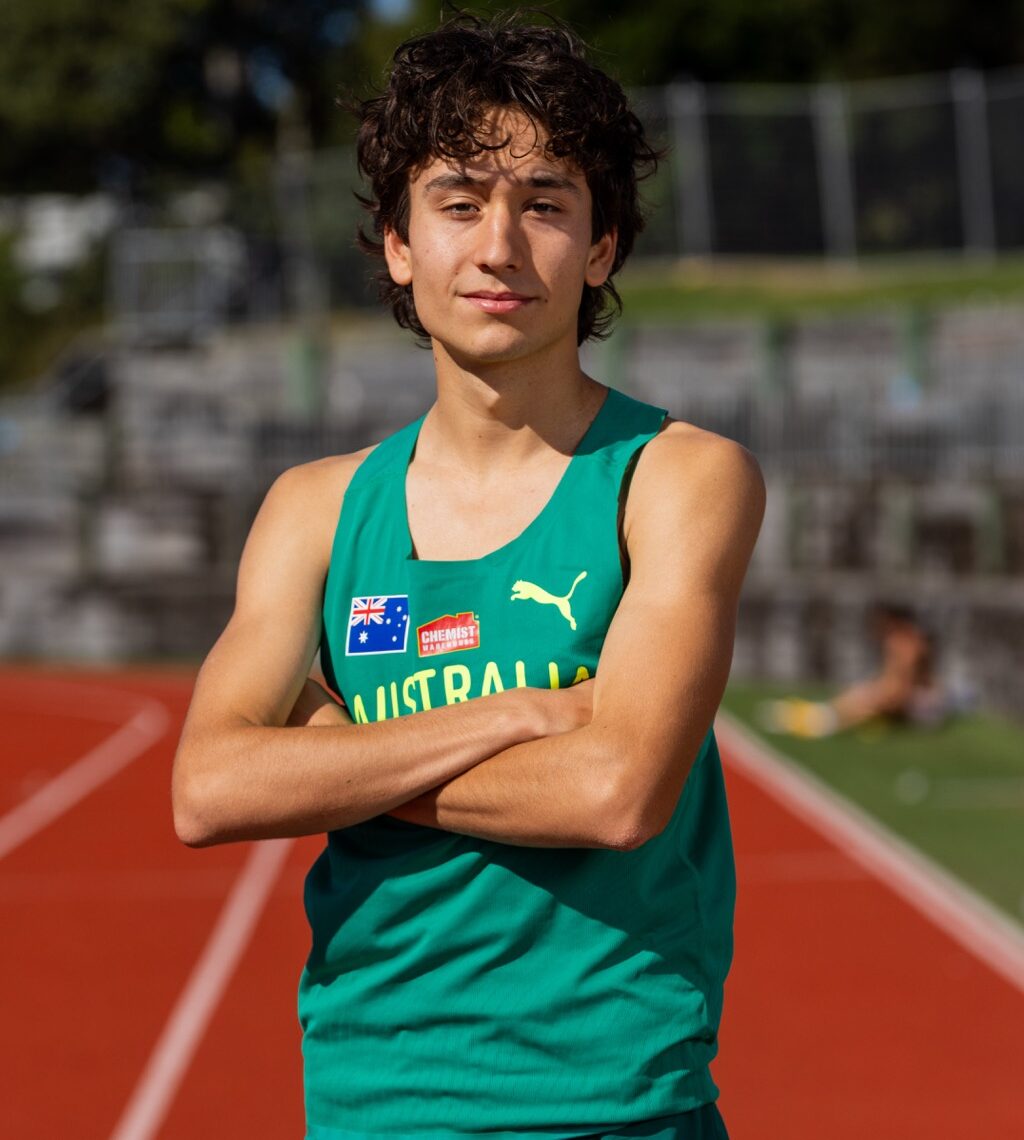 Young male athlete standing with arms crossed on a sunny outdoor running track, wearing a green Australia singlet with sponsor logos.
