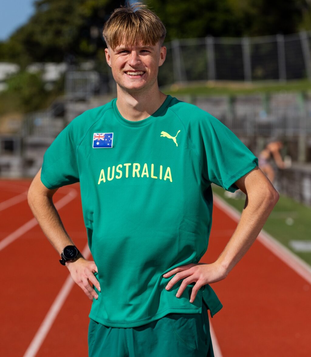 Smiling male athlete on a running track wearing a green Australia jersey with hands on hips.