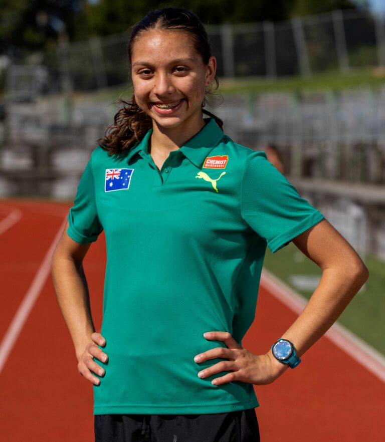 Smiling young female athlete on a running track, wearing a green polo with Australian flag patch and Puma logo, hands on hips.