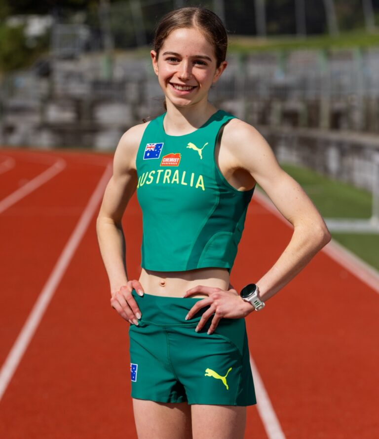 Australian track sprinter posing on a red running track, hands on hips, wearing a green Australia uniform with logos.