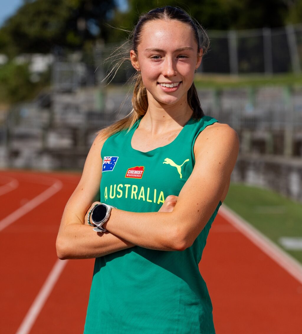 Female Australian track athlete standing with arms crossed on a red running track, smiling at the camera.