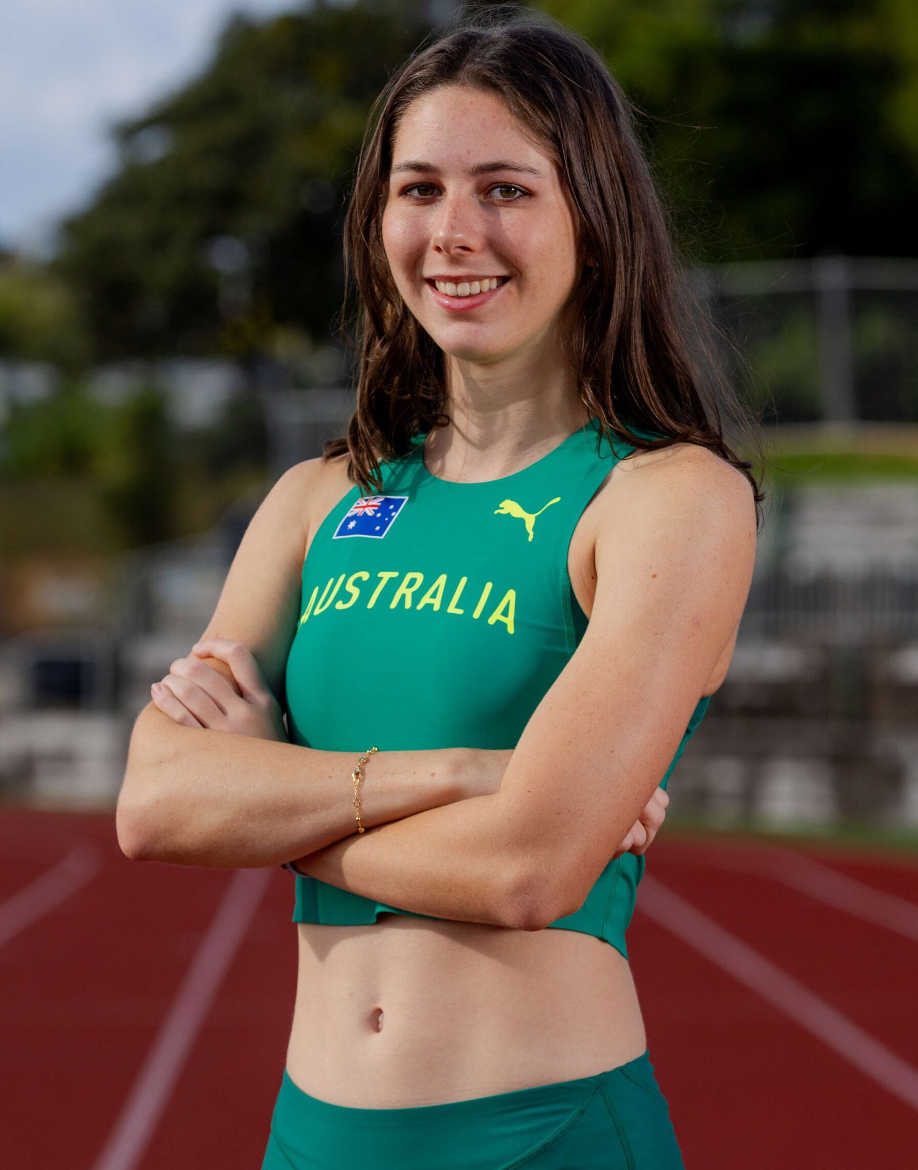 Young female Australian track athlete posing with arms crossed on a red running track, wearing a green athletic top with 'AUSTRALIA' text.
