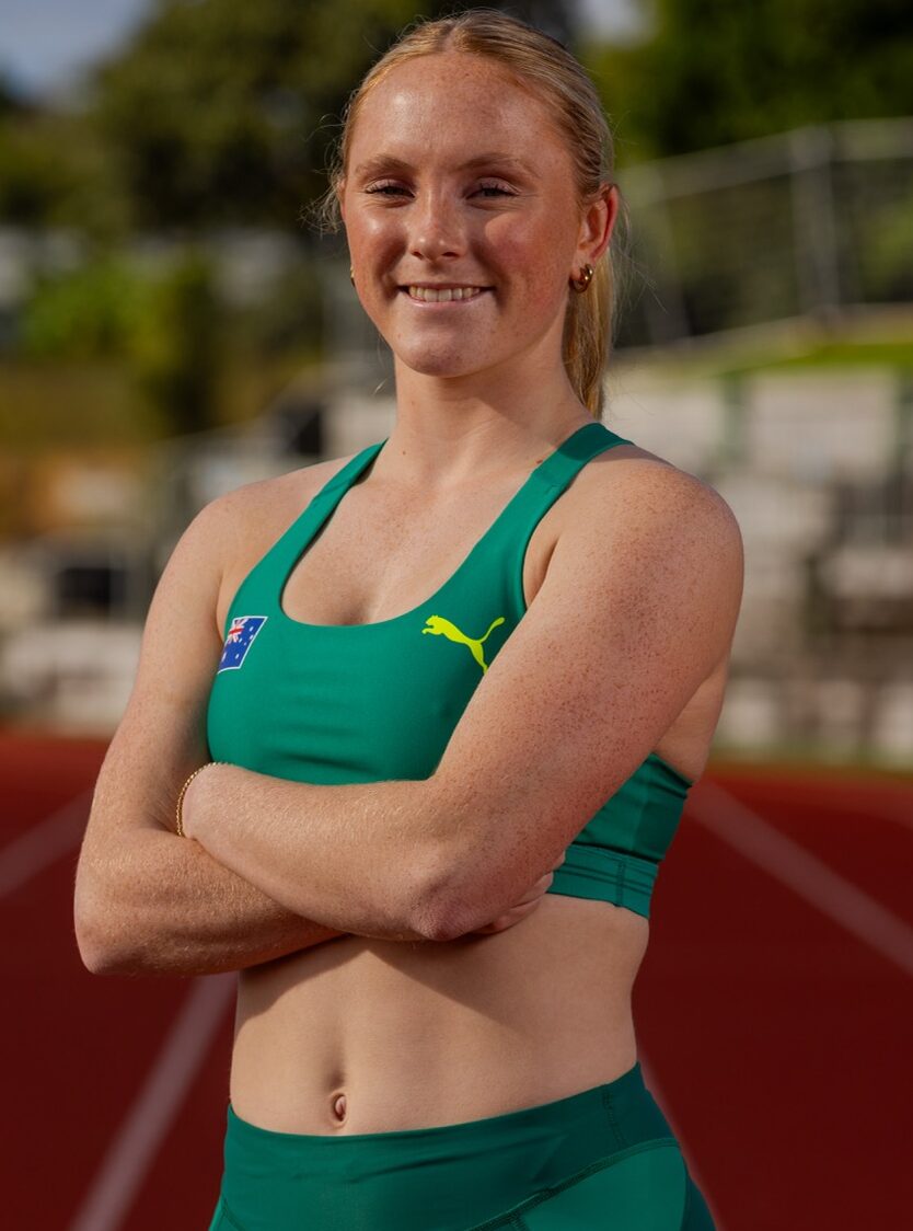 Athletic woman in green sportswear with crossed arms smiling on a red track field, stadium in the background.