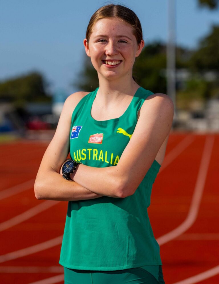 Smiling female athlete in a green Australia uniform standing on a red running track, arms crossed at the front.