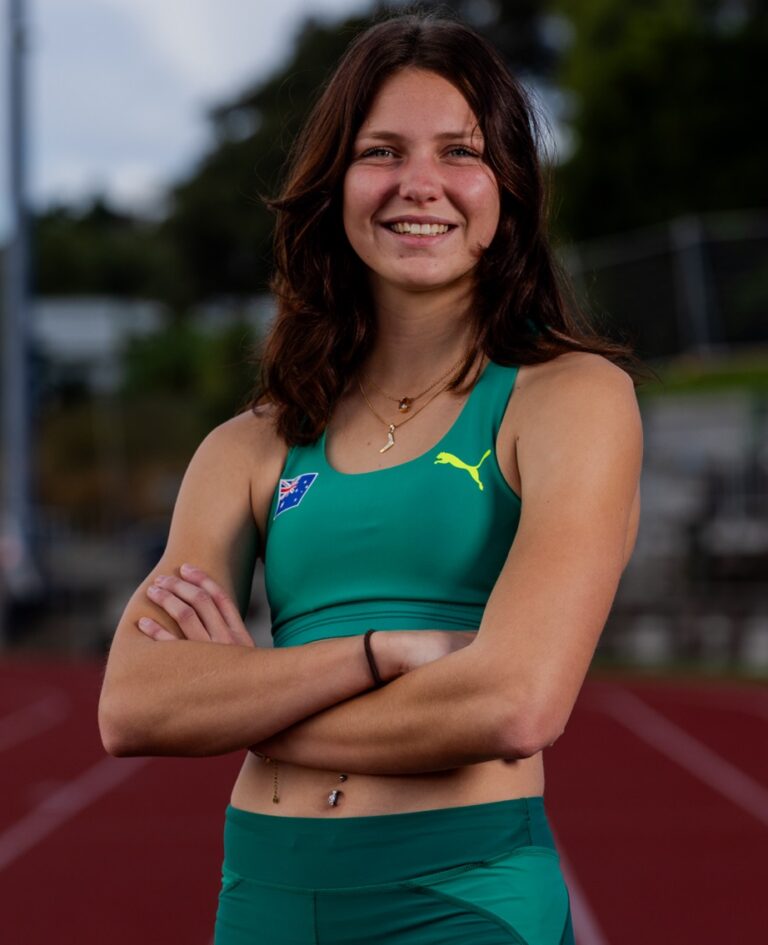 Smiling young woman with brown hair standing with arms crossed on a red running track, wearing teal athletic top and shorts.
