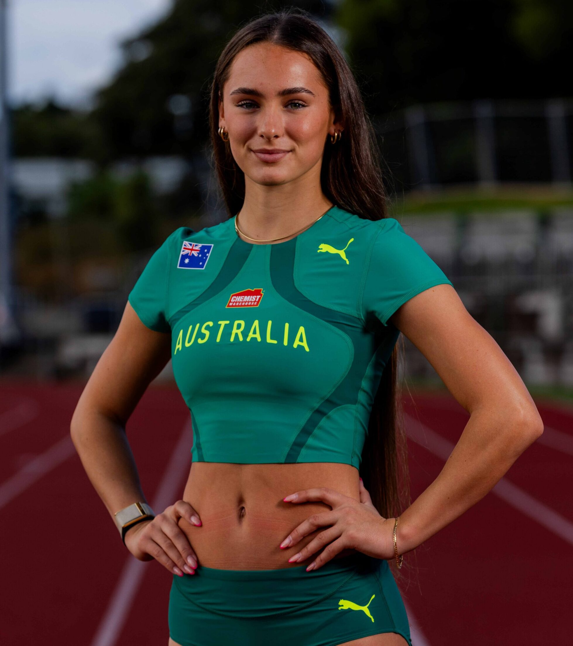 Australian female athlete standing on a track in green sportswear, hands on hips, confident pose.