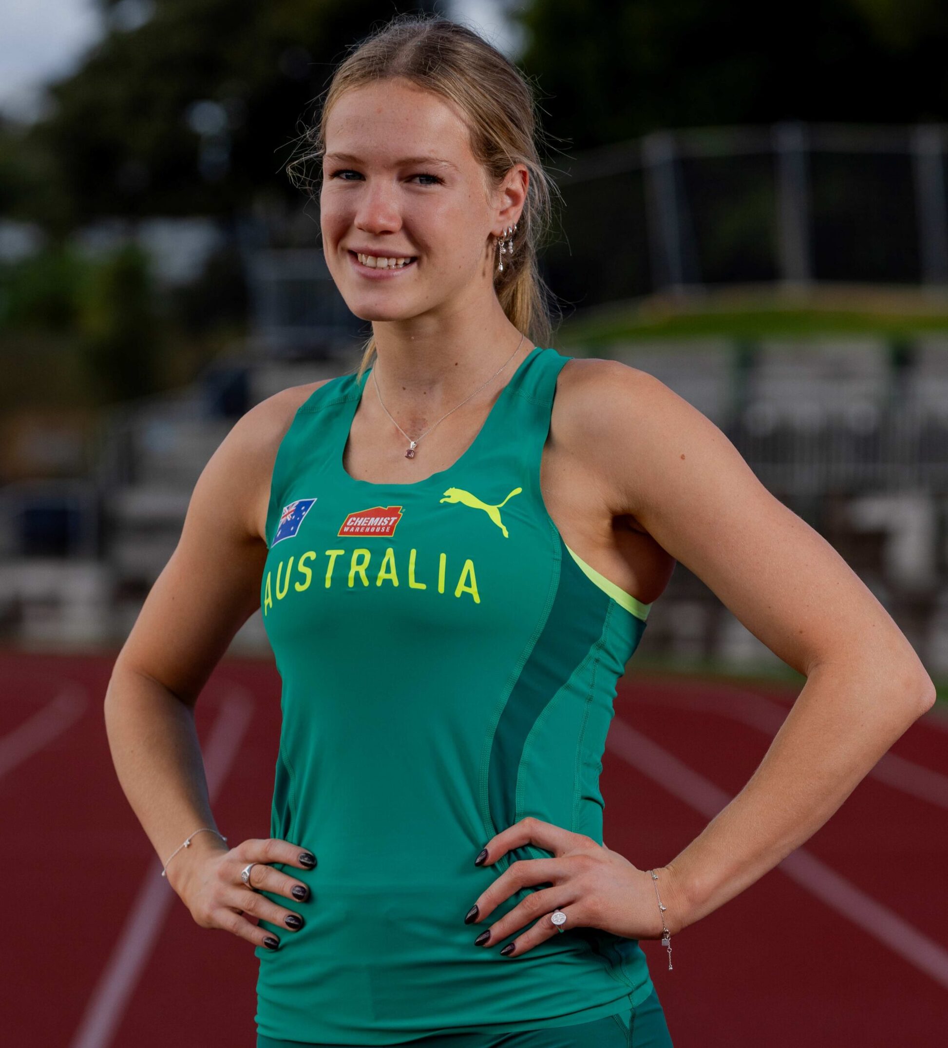 Female athlete in a teal Australia singlet standing with hands on hips on a track field.