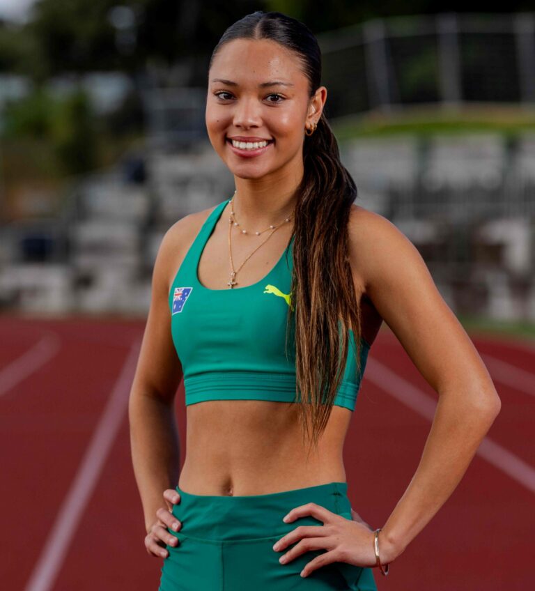 Smiling young woman in teal athletic outfit standing on a running track, hands on hips, outdoors.