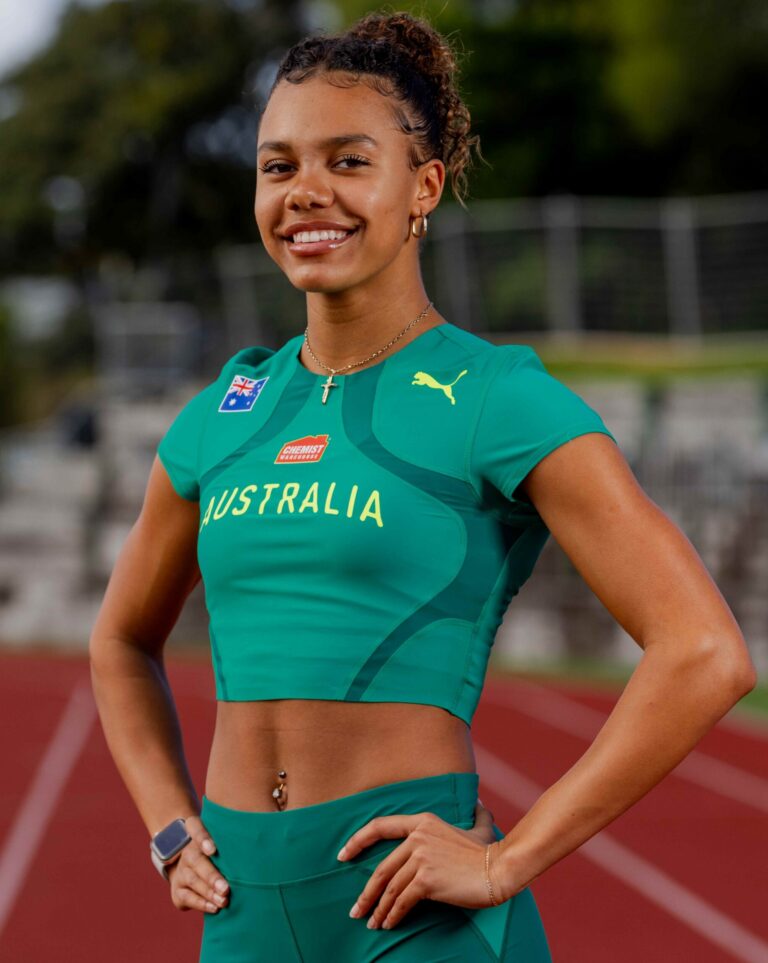 Smiling female athlete in a teal Australia jersey standing on a running track with badges visible on the chest.