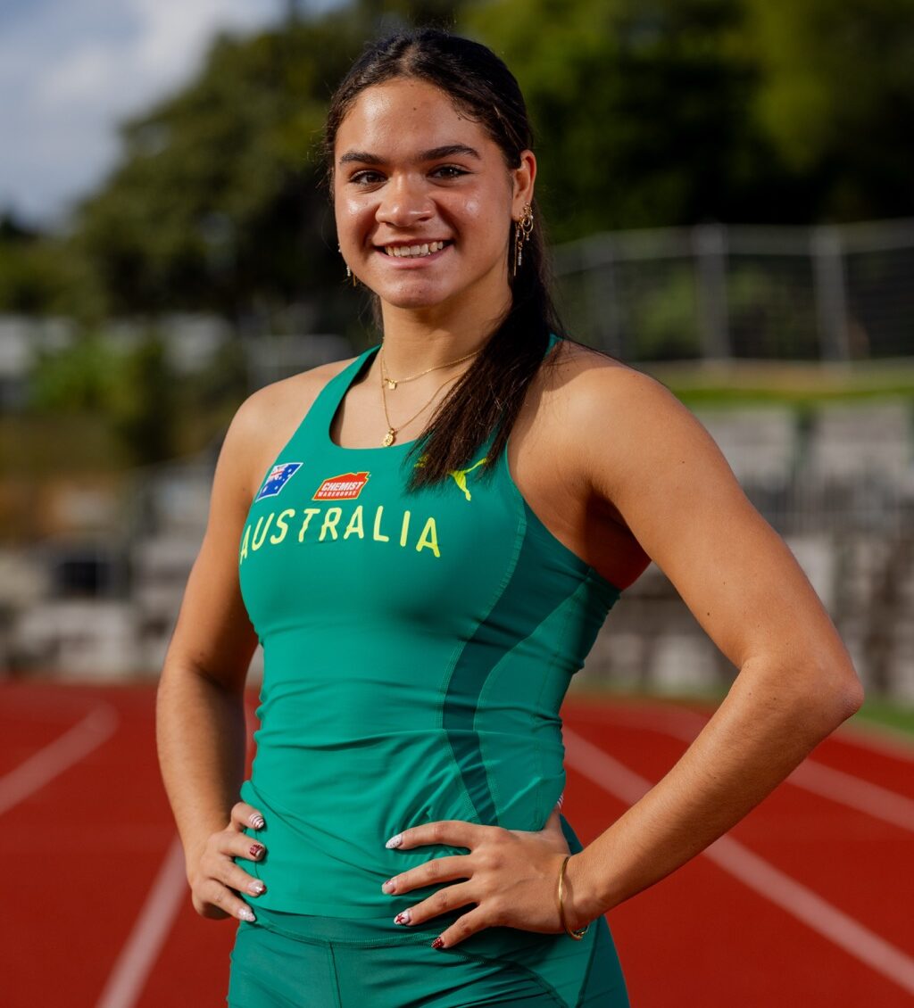 Female athlete in teal Australia track uniform, hands on hips, smiling on a running track at the field edge.