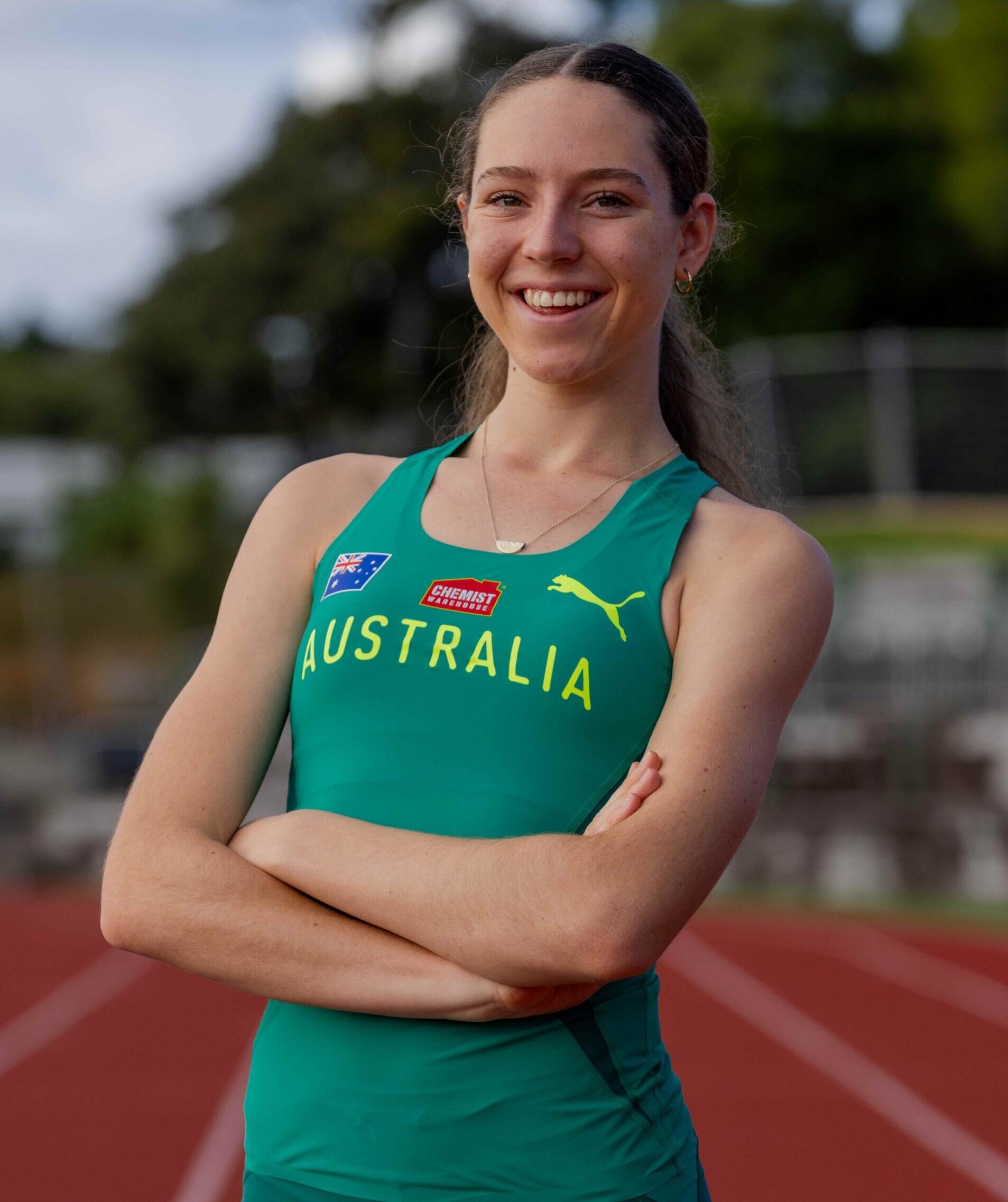 Young woman in teal Australia running kit, arms crossed, smiling on a track with greenery in the background.