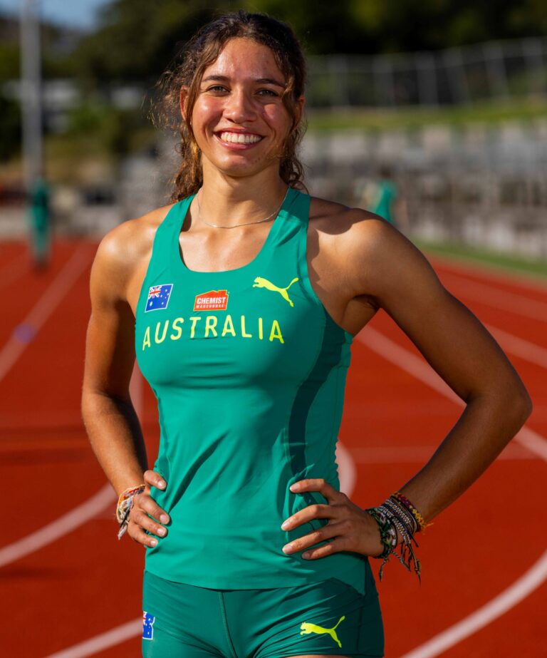 Smiling female Australian track athlete on a running track, hands on hips in a green uniform with Australia text and logos.