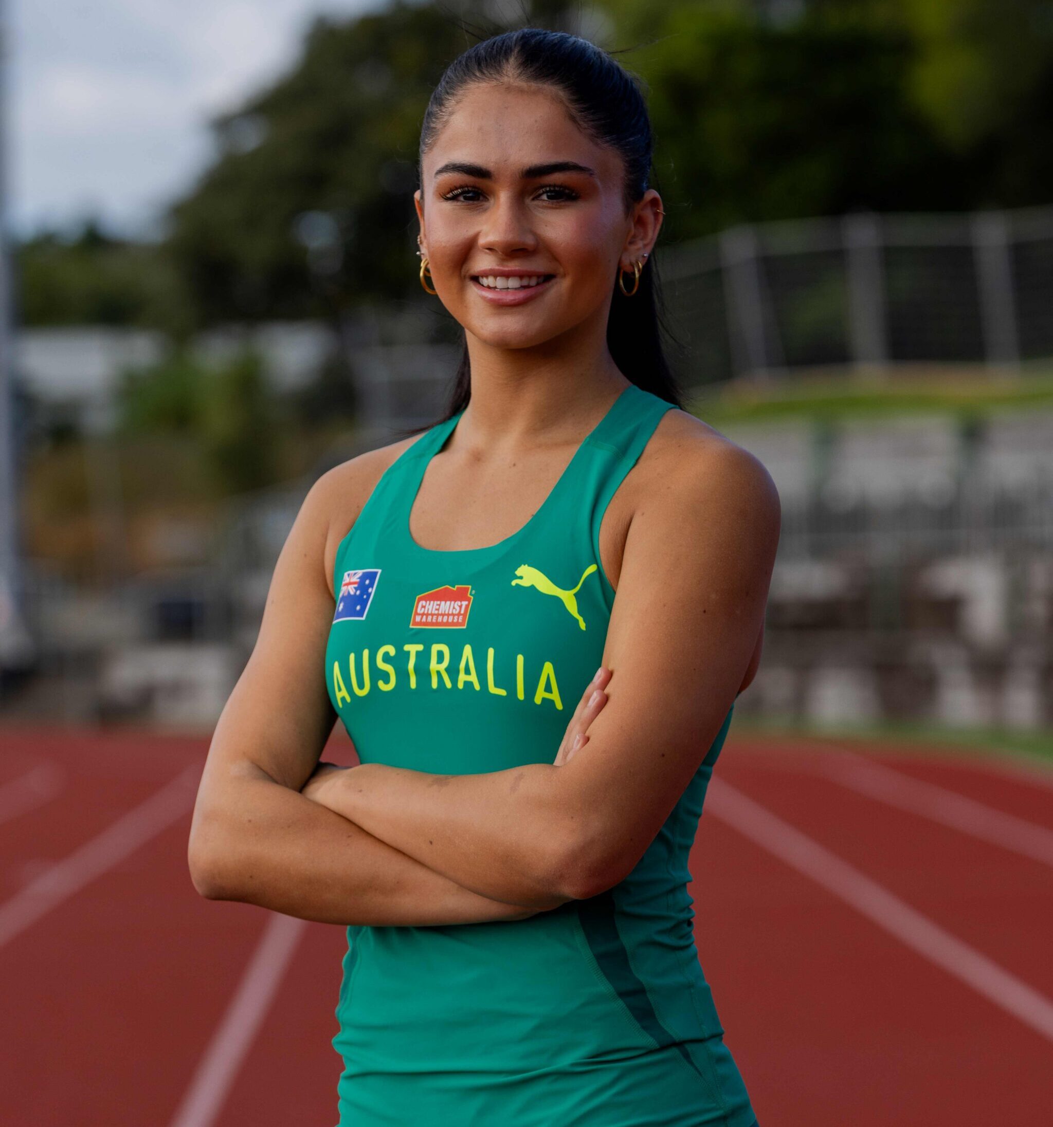 Smiling female athlete in teal Australia uniform with crossed arms on a running track outdoors.