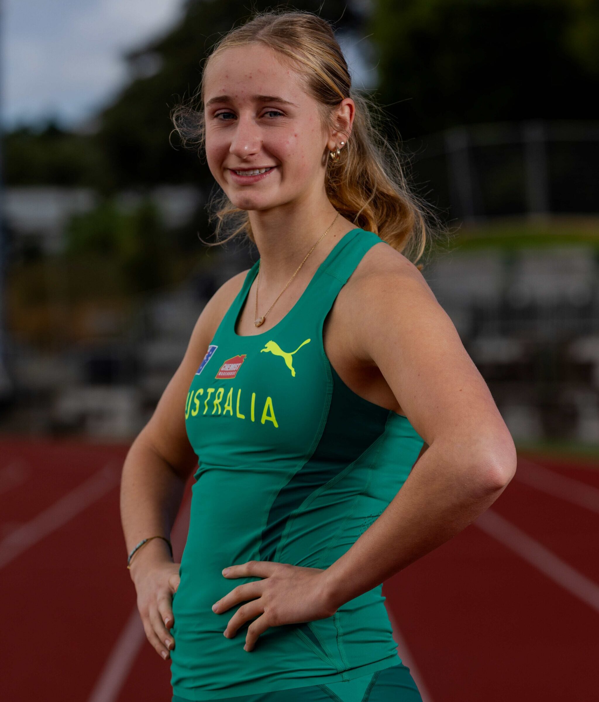 Smiling female athlete in a green Australian track uniform standing on a running track outdoors, hands on hips.