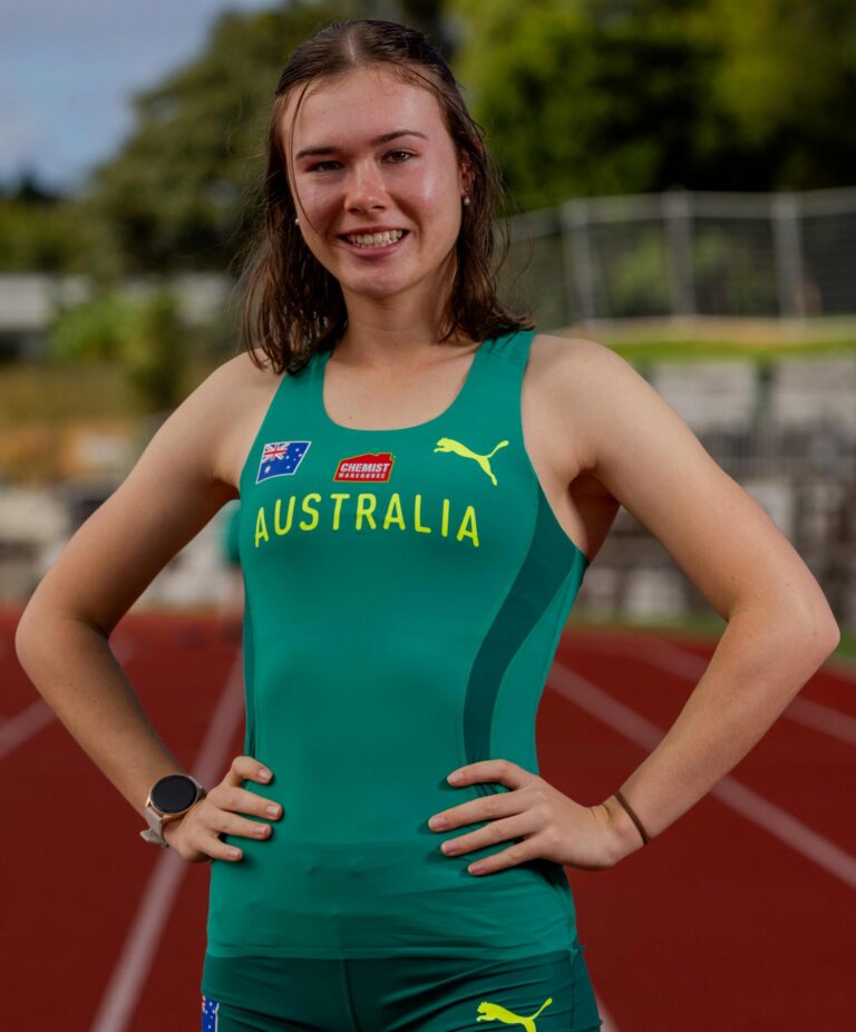 Young female athlete in a green Australia racing suit standing on a red track with hands on hips, smiling at the camera.