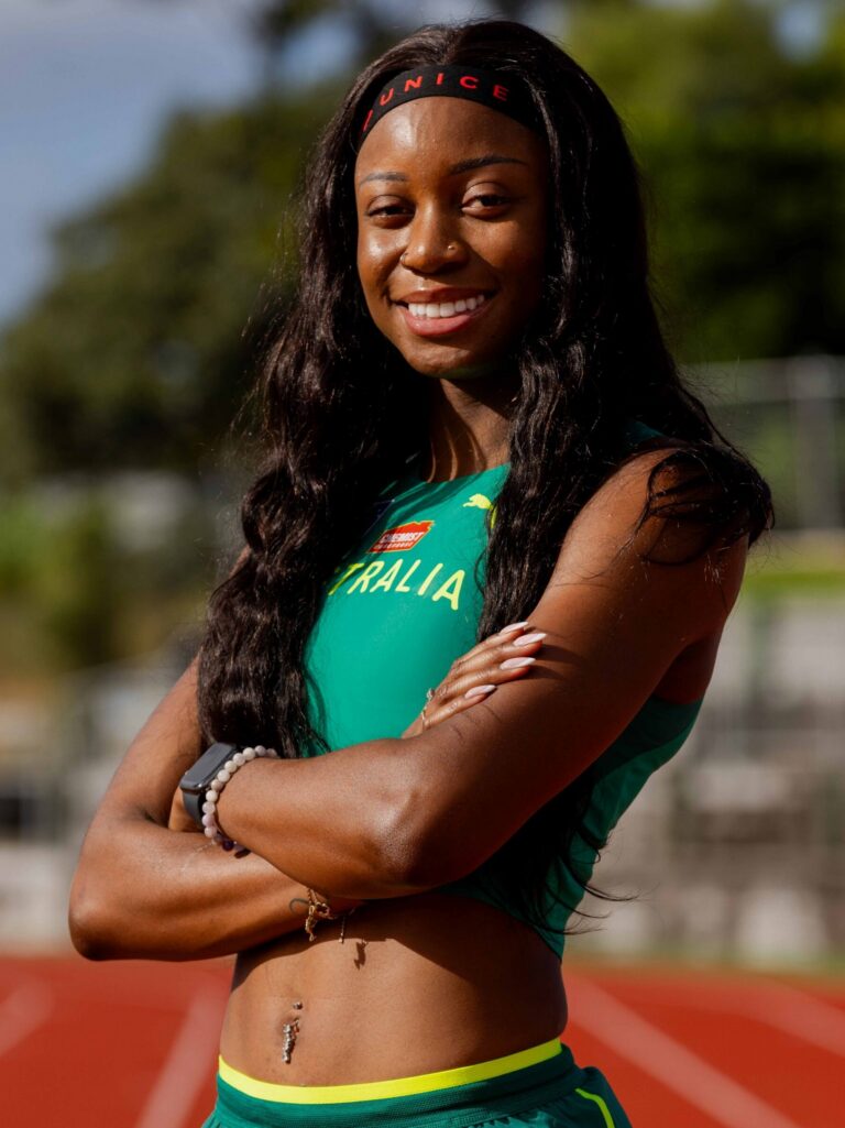 Smiling female athlete in a teal-green sprint uniform, standing with arms crossed on a track field.
