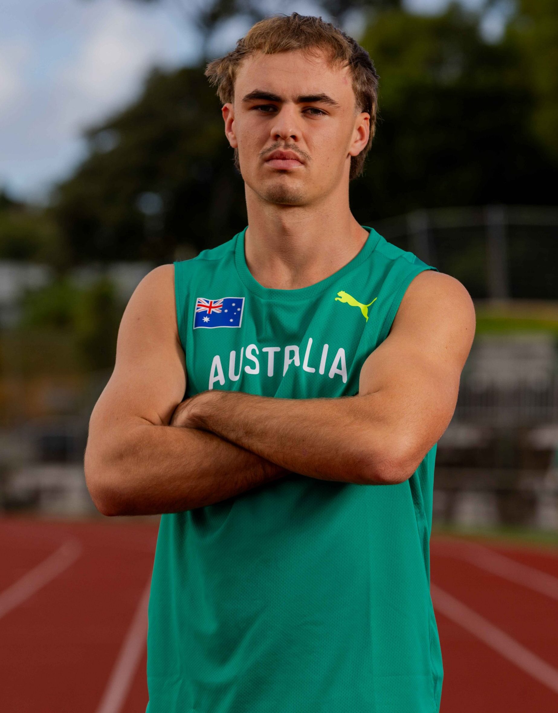 Young male athlete in a green Australia jersey with arms crossed on an outdoor track.