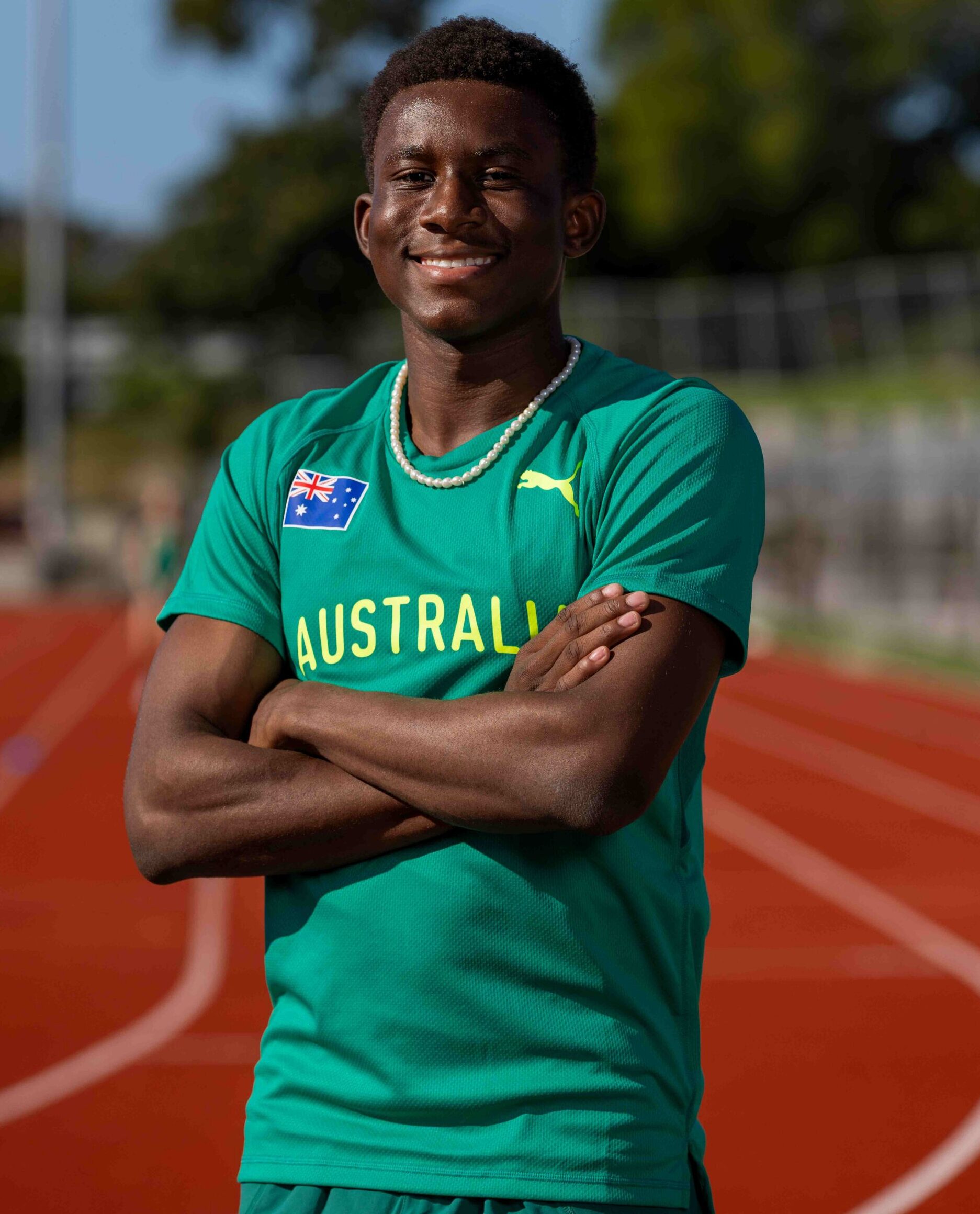 Smiling young athlete in a teal Australian shirt with a pearl necklace, arms crossed on a running track outdoors.