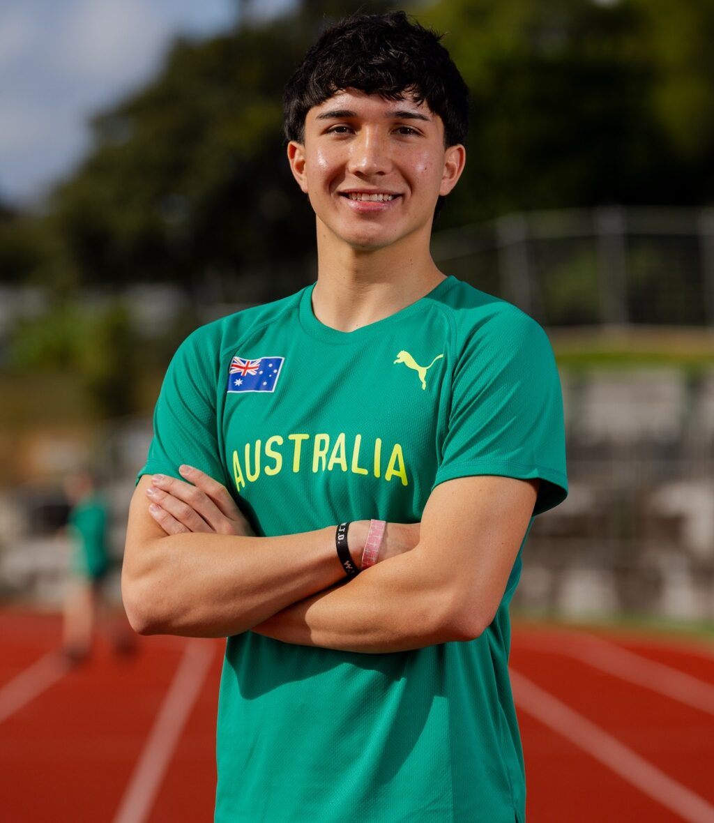 Young man in green Australia athletic jersey with crossed arms on a track field, smiling at camera.