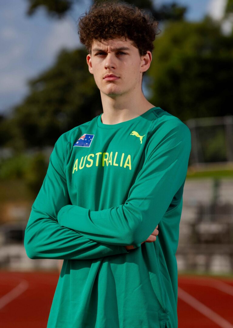 Young man in a green Australia training shirt stands with arms crossed outdoors, looking serious on a track field.