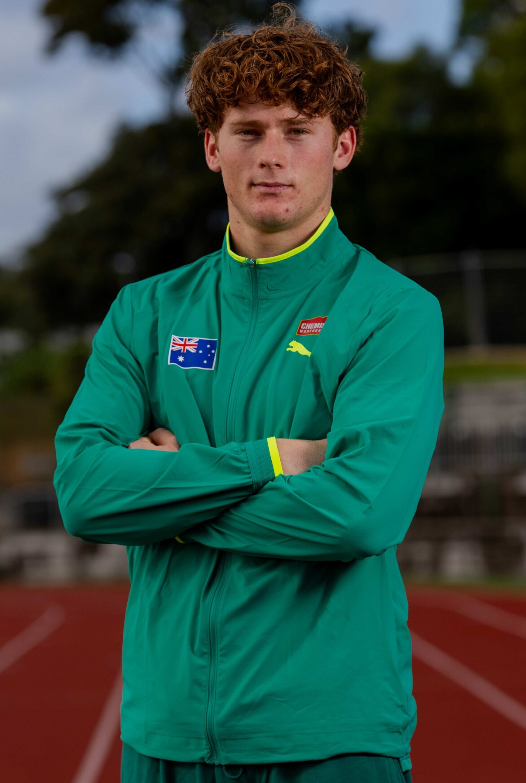 Young man with curly red hair in a green tracksuit, arms crossed, standing on a red running track with lanes visible behind him.