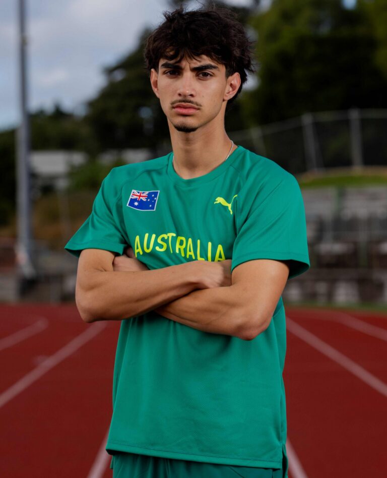Young man in a green Australia jersey with arms crossed on a running track, blurred stadium behind him.