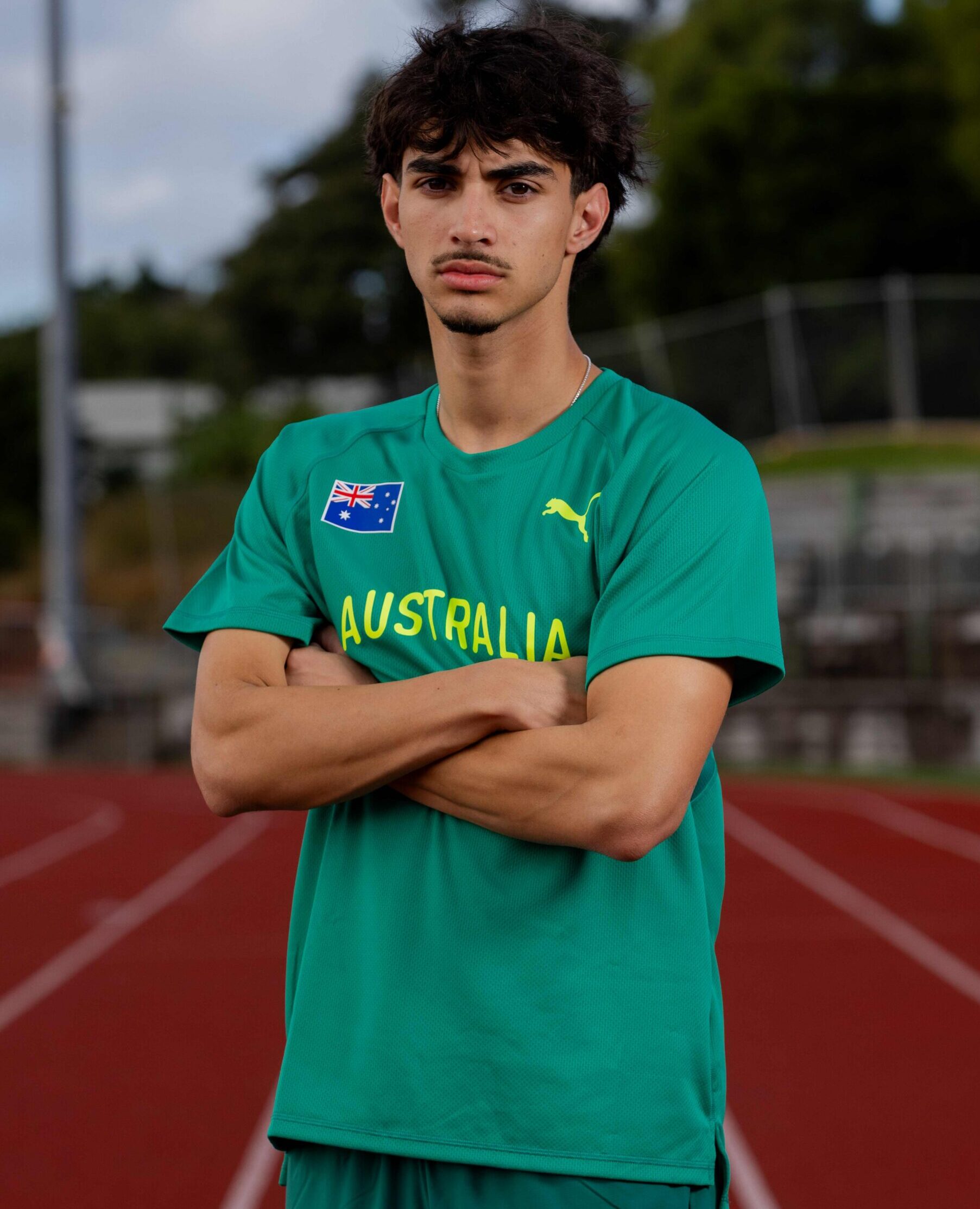Young man in a green Australia jersey with arms crossed on a running track, blurred stadium behind him.