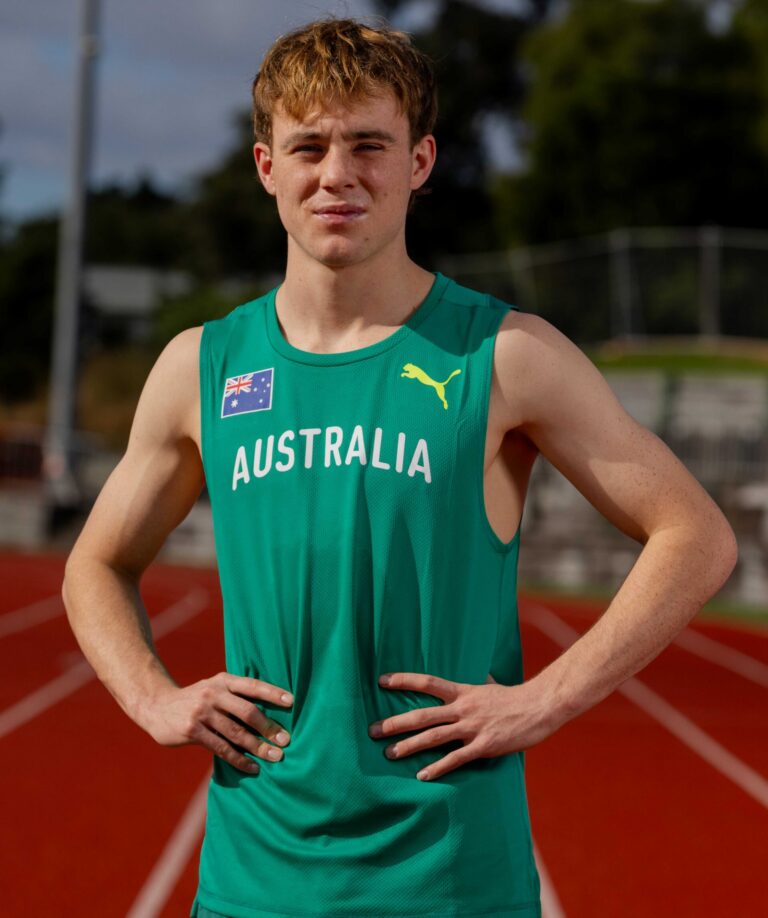 Male athlete in a green Australia singlet stands with hands on hips on a running track; Australian flag emblem visible on chest.